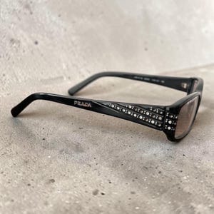 May include: Black Prada sunglasses featuring a rectangular frame and temples embellished with rows of small clear crystals. The brand name Prada is printed in silver lettering on the side of the temple, and the lenses have a dark tint.