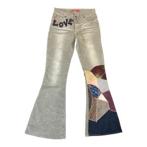 Può includere: Jeans a zampa in velluto a coste grigio chiaro con la scritta "LOVE" a lettere nere sul davanti. I jeans presentano un design patchwork sulla parte inferiore delle gambe con vari tessuti e colori.