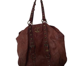 May include: A reddish-brown leather tote bag with a top handle and shoulder straps. The bag features gold-coloured studs and hardware, including buckles and rings. A gold emblem is centered on the front.