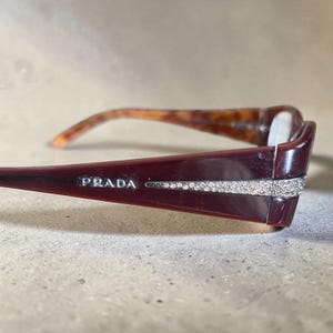 Peut inclure: Lunettes de vue bordeaux foncé avec le logo Prada en lettres argentées sur la branche. La branche présente une incrustation effilée de petits cristaux transparents s'étendant vers la monture avant. L'intérieur de la branche a un motif écaille.
