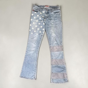 May include: Light wash denim jeans with a flared leg. The left leg features white star accents, while the right leg has gray patchwork. The jeans have a classic five-pocket design and a red logo tag.