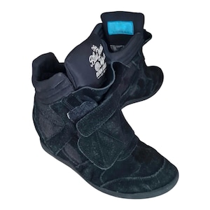 May include: Black high-top sneakers with a wedge heel. The sneakers feature a suede-like texture, a velcro strap, and the text "The Blue Jeans" on the tongue. The shoes have a padded collar and tongue.