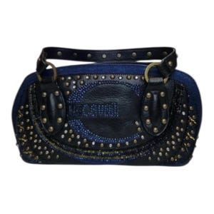 May include: A dark blue denim handbag with black leather details and gold-coloured studs. The bag features the text "JUST CAVALLI" and a black handle. The bag is embellished with small, sparkling rhinestones.