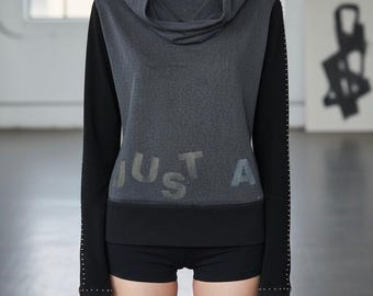 May include: A grey hooded sweatshirt with black sleeves and shorts. The sweatshirt has the text "JUST A" in a faded, metallic print. Small silver studs are on the sleeves.