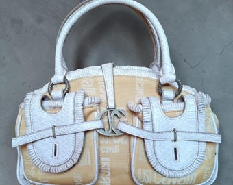 May include: A cream and tan handbag with a top handle and two smaller handles. The bag features a repeating pattern of the words "Just Cavalli" and has white accents, including the handle, trim, and decorative elements. A silver-tone clasp is centered on the front.