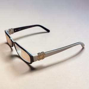 Peut inclure: Lunettes de soleil rectangulaires dotées d'une monture bicolore avec une face brun foncé et des branches couleur crème. Les charnières sont ornées d'un emblème métallique doré en forme de fleur. Les verres ont une teinte ambrée claire.