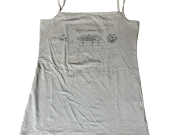 May include: A light beige camisole top with thin spaghetti straps. The top features a vintage-style print with floral and script elements. The text includes the words "COST COPINS" and other script. The fabric appears to be soft and slightly wrinkled.