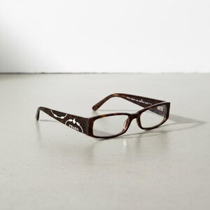 Peut inclure: Lunettes de vue rectangulaires à motif écaille de tortue avec des verres transparents. Les branches présentent un logo Prada blanc avec un motif circulaire stylisé. La monture est en acétate marbré de brun et de noir.