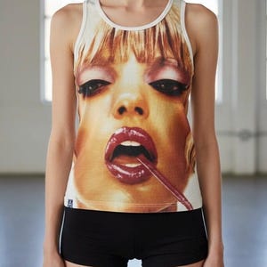 May include: A white tank top with a large, close-up graphic of a woman's face with red lipstick and a lollipop. The model is wearing black shorts. The top has a white trim.
