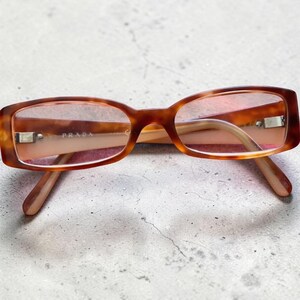 Peut inclure: Lunettes rectangulaires avec une monture à motif écaille de tortue marron et des verres teintés rose clair. La branche intérieure de la monture porte le nom de marque PRADA imprimé en lettres blanches. Les lunettes ont des branches fines assorties.