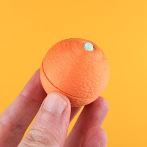 Peut inclure: Une sphère orange avec un accent bleu clair sur le dessus. La sphère est tenue dans une main sur un fond jaune vif. La sphère semble être faite d'un matériau texturé.