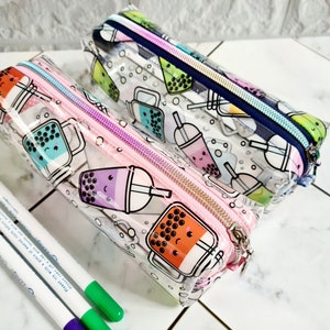 May include: Two clear plastic pencil cases with zipper closures. The cases are decorated with a pattern of cartoon bubble tea cups. One case is pink and the other is blue.