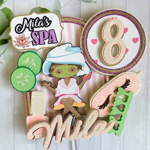 Puede incluir: Adorno para tarta con tem&aacute;tica de spa. Presenta una ni&ntilde;a de dibujos animados con mascarilla, rodajas de pepino y el texto "Mila's SPA". Tambi&eacute;n incluye el n&uacute;mero "8", junto con detalles de purpurina rosa y dorada.