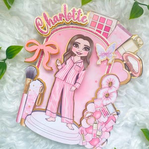 Personalized makeup-themed cake topper featuring a girl in pink striped pajamas surrounded by beauty and spa accessories including makeup brushes, lipstick, blusher, eyeshadow, skincare items, and a pink bow. The topper is decorated in pink glitter card and gold mirror  with the name and age of the birthday girl, making it perfect for a glam birthday, spa party, or makeup-themed celebration.
