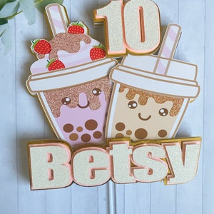 May include: A layered cake topper featuring two cartoon bubble tea cups with smiling faces. One cup is pink with strawberry topping, the other is brown. The number 10 and the name "Betsy" are also included, all with glitter accents.