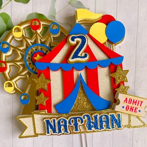 May include: A colorful circus-themed cake topper featuring a Ferris wheel, a red and white striped tent, and the name "NATHAN" in blue glitter. Includes a ticket that says "ADMIT ONE" and the number "2".