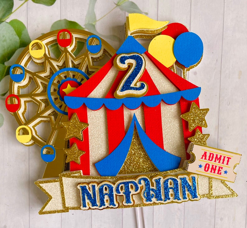 May include: A colorful circus-themed cake topper featuring a Ferris wheel, a red and white striped tent, and the name "NATHAN" in blue glitter. Includes a ticket that says "ADMIT ONE" and the number "2".