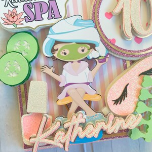 Puede incluir: Adorno para tarta con tem&aacute;tica de spa que presenta a una ni&ntilde;a de dibujos animados con mascarilla, pepinos y esmalte de u&ntilde;as. El adorno incluye el texto "Katherine's SPA" y el n&uacute;mero 10, con detalles en rosa y dorado.