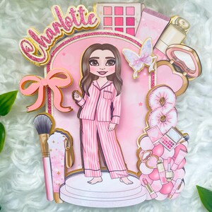 Personalized makeup-themed cake topper featuring a girl in pink striped pajamas surrounded by beauty and spa accessories including makeup brushes, lipstick, blusher, eyeshadow, skincare items, and a pink bow. The topper is decorated in pink glitter card and gold mirror  with the name and age of the birthday girl, making it perfect for a glam birthday, spa party, or makeup-themed celebration.