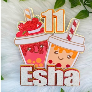 May include: A vibrant cake topper with two bubble tea cups, one pink and one orange, each with a straw. The number "11" and the name "Esha" are included. The topper is decorated with glitter and strawberries.