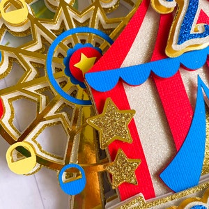 May include: A colorful, layered paper craft featuring a Ferris wheel, circus tent, and gold glitter stars. The Ferris wheel has red, blue, and yellow gondolas. The circus tent is red and white with blue trim. The number "2" is visible.