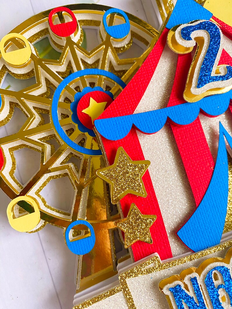 May include: A colorful, layered paper craft featuring a Ferris wheel, circus tent, and gold glitter stars. The Ferris wheel has red, blue, and yellow gondolas. The circus tent is red and white with blue trim. The number "2" is visible.