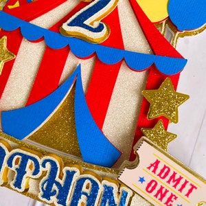 May include: A colorful circus-themed cake topper with a red and white striped tent, blue accents, and gold glitter details. Features a blue number 2, a ticket that says "ADMIT ONE", and the name "NATHAN" in blue glitter letters. Includes yellow and blue balloons.