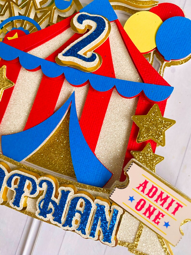 May include: A colorful circus-themed cake topper with a red and white striped tent, blue accents, and gold glitter details. Features a blue number 2, a ticket that says "ADMIT ONE", and the name "NATHAN" in blue glitter letters. Includes yellow and blue balloons.