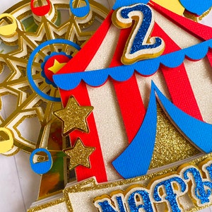 May include: A colorful circus-themed cake topper featuring a Ferris wheel, a circus tent, and the number "2". The topper includes gold glitter accents, red, blue, and yellow details, and a banner with a name.