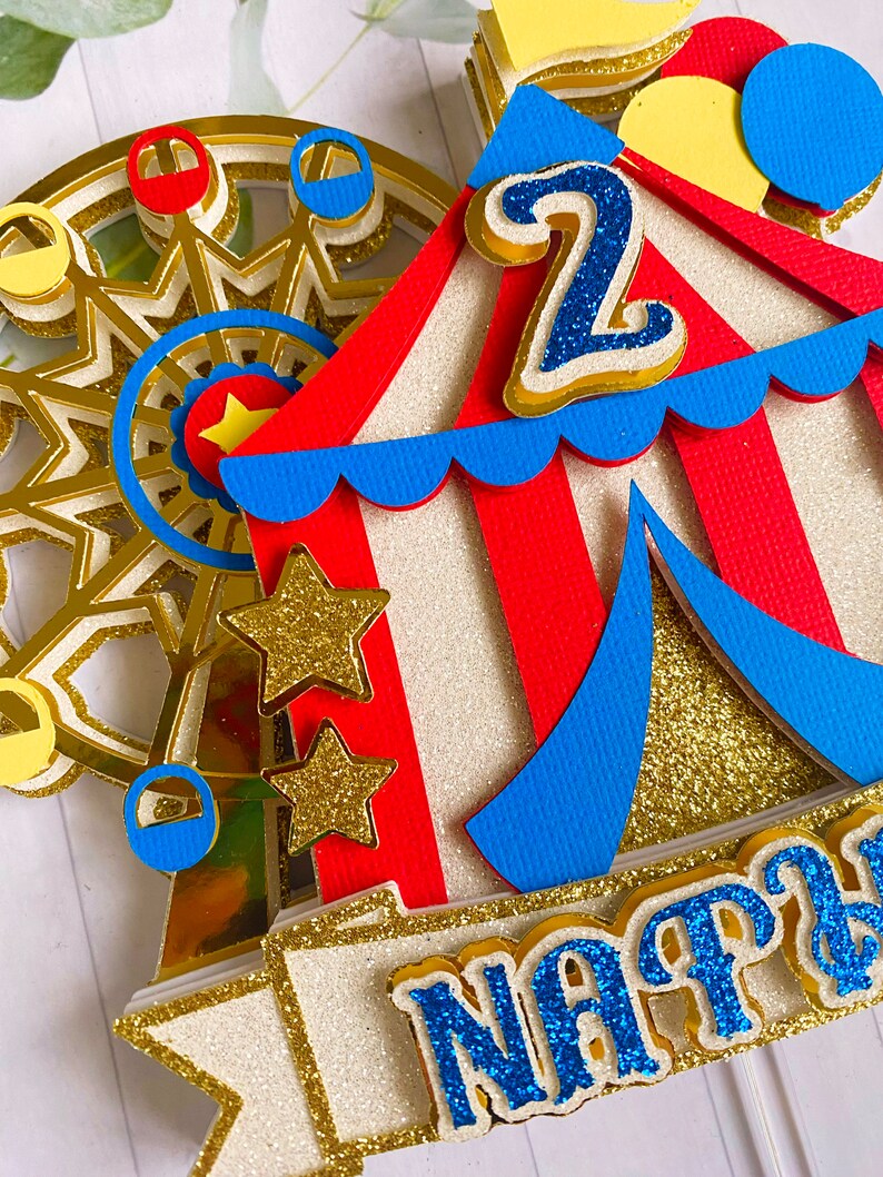 May include: A colorful circus-themed cake topper featuring a Ferris wheel, a circus tent, and the number "2". The topper includes gold glitter accents, red, blue, and yellow details, and a banner with a name.