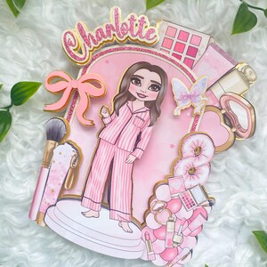 Personalized makeup-themed cake topper featuring a girl in pink striped pajamas surrounded by beauty and spa accessories including makeup brushes, lipstick, blusher, eyeshadow, skincare items, and a pink bow. The topper is decorated in pink glitter card and gold mirror  with the name and age of the birthday girl, making it perfect for a glam birthday, spa party, or makeup-themed celebration.