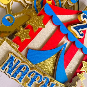 May include: A multi-layered circus-themed cake topper with a Ferris wheel, tent, and stars. The topper features red and white stripes, gold glitter accents, and the name "Nathan" in blue glitter. A ticket that says "Admit One" is also visible.