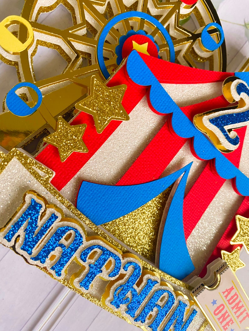 May include: A multi-layered circus-themed cake topper with a Ferris wheel, tent, and stars. The topper features red and white stripes, gold glitter accents, and the name "Nathan" in blue glitter. A ticket that says "Admit One" is also visible.