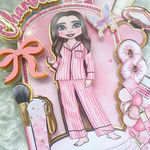 Personalized makeup-themed cake topper featuring a girl in pink striped pajamas surrounded by beauty and spa accessories including makeup brushes, lipstick, blusher, eyeshadow, skincare items, and a pink bow. The topper is decorated in pink glitter card and gold mirror  with the name and age of the birthday girl, making it perfect for a glam birthday, spa party, or makeup-themed celebration.