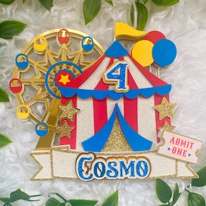 May include: A colorful circus-themed cake topper with a Ferris wheel, a tent, and balloons. The number 4 is displayed, along with the name "Cosmo" and an "Admit One" ticket. Gold glitter accents the design.