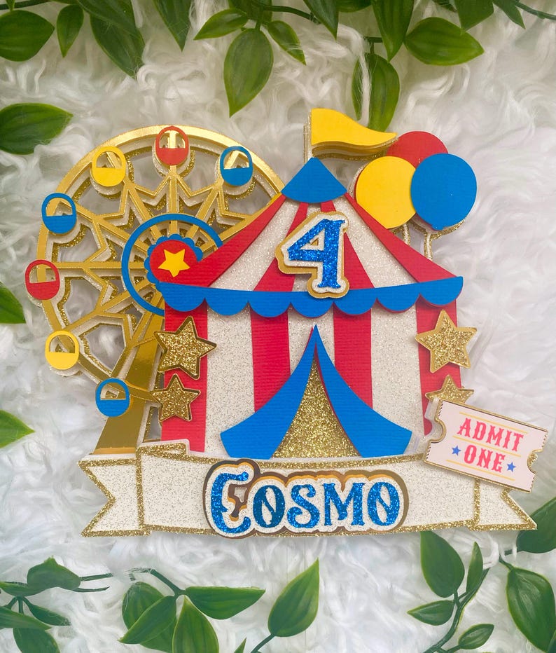 May include: A colorful circus-themed cake topper with a Ferris wheel, a tent, and balloons. The number 4 is displayed, along with the name "Cosmo" and an "Admit One" ticket. Gold glitter accents the design.