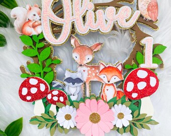 Op de afbeelding: Een taarttopper met bos thema met de naam "Olive" in roze glitterletters. Het ontwerp bevat een eekhoorn, hert, vos, wasbeer en uil, samen met paddenstoelen, bloemen en het cijfer "1", geschikt voor een eerste verjaardag.