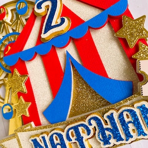 May include: A colorful circus-themed cake topper with a red and white striped tent, blue accents, and gold glitter stars. The number "2" and the name "NATHAN" are prominently displayed in blue glitter.