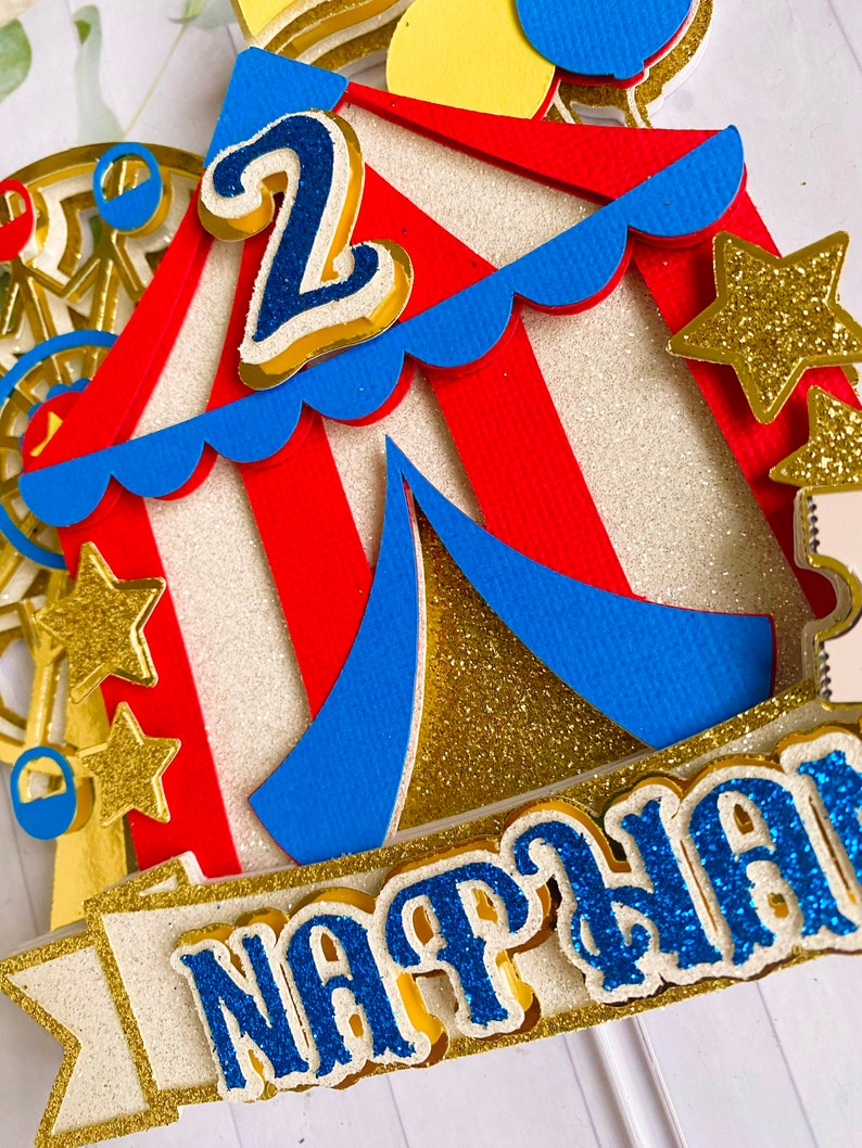 May include: A colorful circus-themed cake topper with a red and white striped tent, blue accents, and gold glitter stars. The number "2" and the name "NATHAN" are prominently displayed in blue glitter.