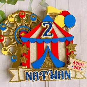 May include: A festive circus-themed cake topper featuring a Ferris wheel, a red and white striped tent, and the name "NATHAN". The number "2" is prominently displayed, along with gold stars, balloons, and an "ADMIT ONE" ticket. The design incorporates gold glitter accents.