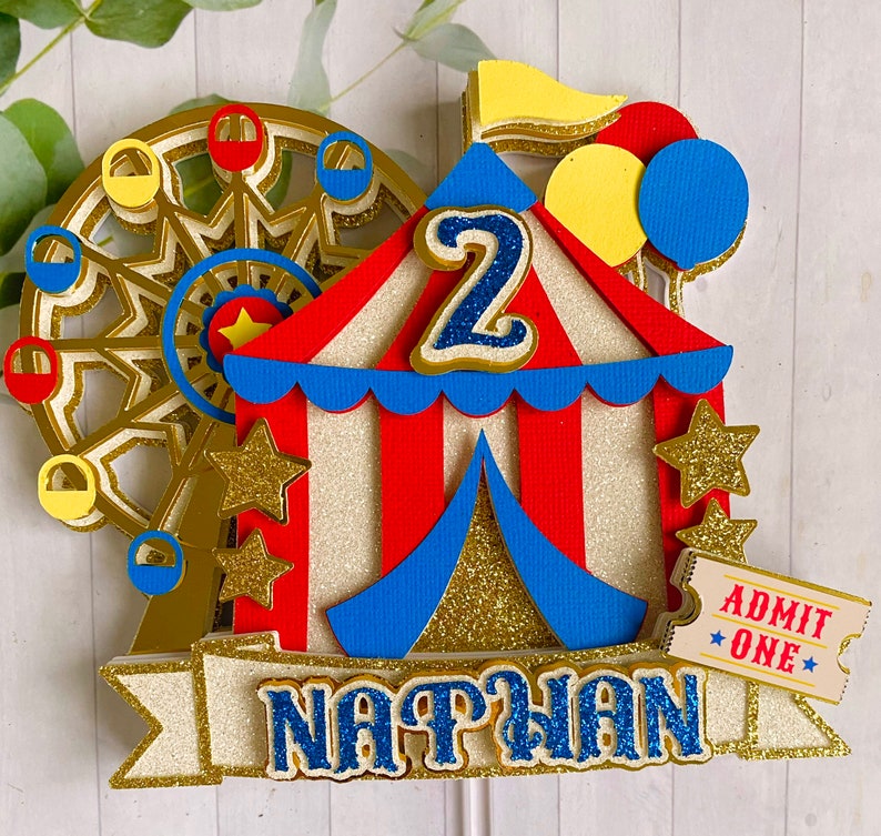 May include: A festive circus-themed cake topper featuring a Ferris wheel, a red and white striped tent, and the name "NATHAN". The number "2" is prominently displayed, along with gold stars, balloons, and an "ADMIT ONE" ticket. The design incorporates gold glitter accents.