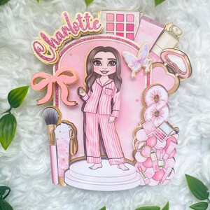 Personalized makeup-themed cake topper featuring a girl in pink striped pajamas surrounded by beauty and spa accessories including makeup brushes, lipstick, blusher, eyeshadow, skincare items, and a pink bow. The topper is decorated in pink glitter card and gold mirror  with the name and age of the birthday girl, making it perfect for a glam birthday, spa party, or makeup-themed celebration.