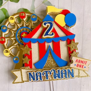May include: A colorful circus-themed cake topper featuring a Ferris wheel, a red and white striped tent, and gold stars. The topper includes the name "NATHAN" and the number "2" in blue glitter. A ticket that says "ADMIT ONE" is also included.