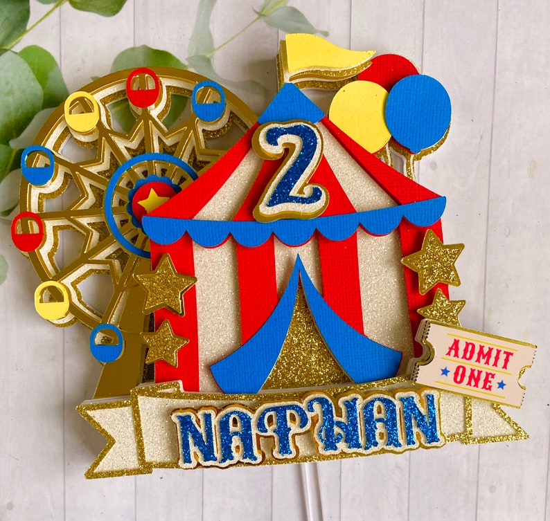 May include: A colorful circus-themed cake topper featuring a Ferris wheel, a red and white striped tent, and gold stars. The topper includes the name "NATHAN" and the number "2" in blue glitter. A ticket that says "ADMIT ONE" is also included.