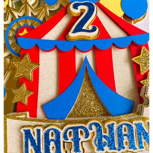 May include: A colorful circus-themed cake topper with a red and white striped tent, blue accents, and gold glitter stars. The number "2" is prominently displayed, along with the name "Nathan" in blue glitter. A Ferris wheel is visible in the background.