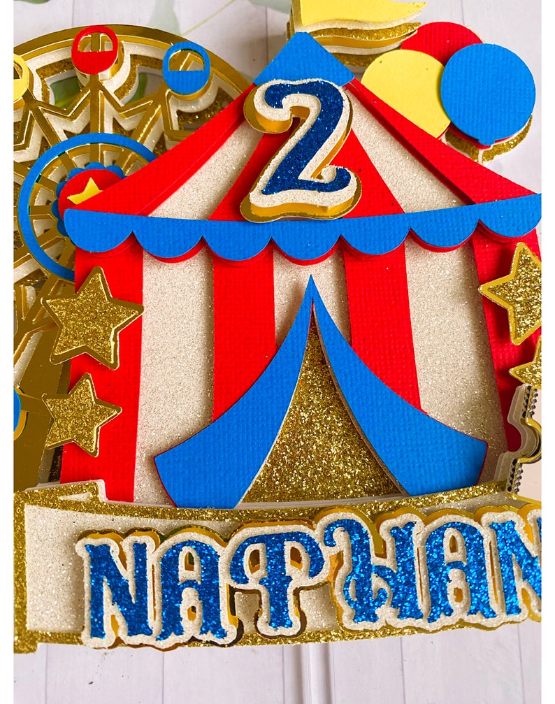 May include: A colorful circus-themed cake topper with a red and white striped tent, blue accents, and gold glitter stars. The number "2" is prominently displayed, along with the name "Nathan" in blue glitter. A Ferris wheel is visible in the background.