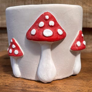 May include: A white ceramic plant pot decorated with three red mushroom designs. Each mushroom features white spots. The pot is cylindrical and rests on a wooden surface.