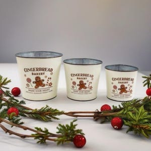 May include: Three cream-coloured metal buckets with a distressed finish, featuring the text "Gingerbread Bakery" and a gingerbread man graphic. The buckets are arranged in a row, with a garland and red ornaments.