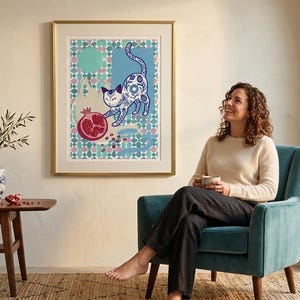 Puede incluir: Una impresión artística enmarcada que presenta un gato estilizado con patrones azules y blancos, interactuando con una granada. La obra de arte cuelga de una pared color crema sobre una mesa de madera con un jarrón. Una mujer está sentada en un sillón verde azulado.