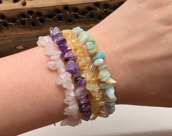 May include: A set of four gemstone bracelets, each featuring a different color of chipped stones. The bracelets are stacked on a person's wrist, starting with a light pink bracelet, followed by a purple bracelet, then a yellow bracelet, and finally a light blue bracelet.